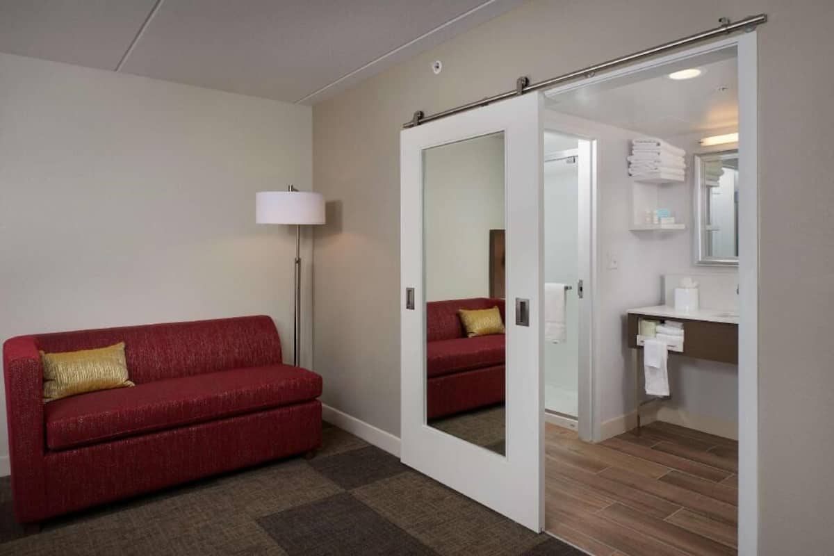 A hotel room with a red sofa, a mirror, a lamp, and a bathroom visible through an open door.