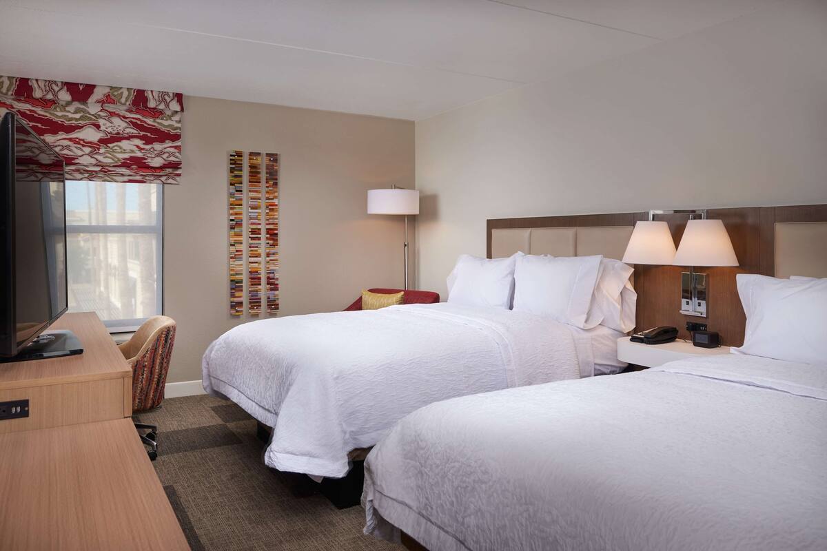 A hotel room with two beds, a desk, a television, a lamp, and a wall with a patterned curtain.