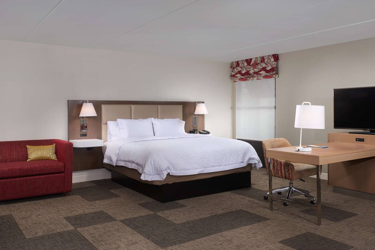 A hotel room with a bed, a desk, a TV, a red sofa, and a chair.