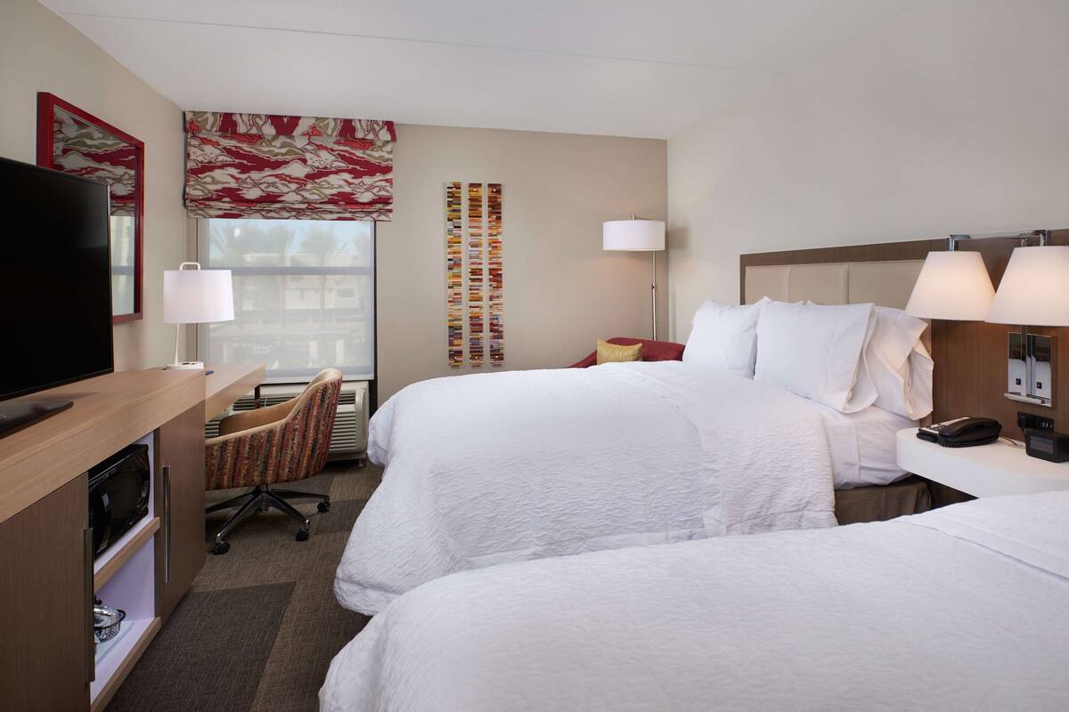 A hotel room with two beds, a desk with a flat-screen TV, a chair, a lamp, and a decorative wall panel.