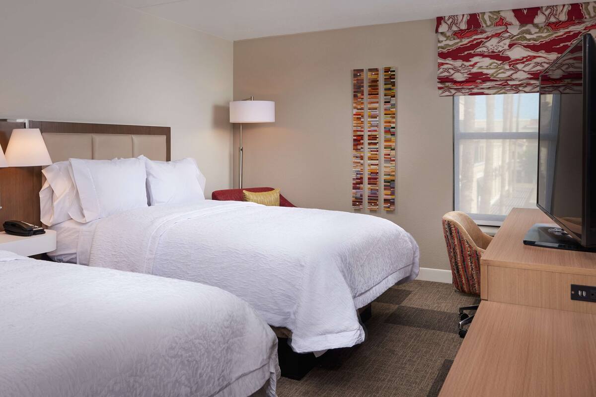 A hotel room with two beds, a desk, a chair, a lamp, and a TV.