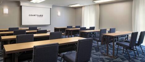 Ein Konferenzraum mit mehreren Tischen und Stühlen, einer Projektionsleinwand mit der Aufschrift "COURTYARD BY MARIEOT" und einem Teppichboden.