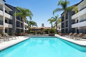 A swimming pool with lounge chairs and palm trees, surrounded by multi-story buildings with balconies. - Courtyard by Marriott Fresno (Fresno)