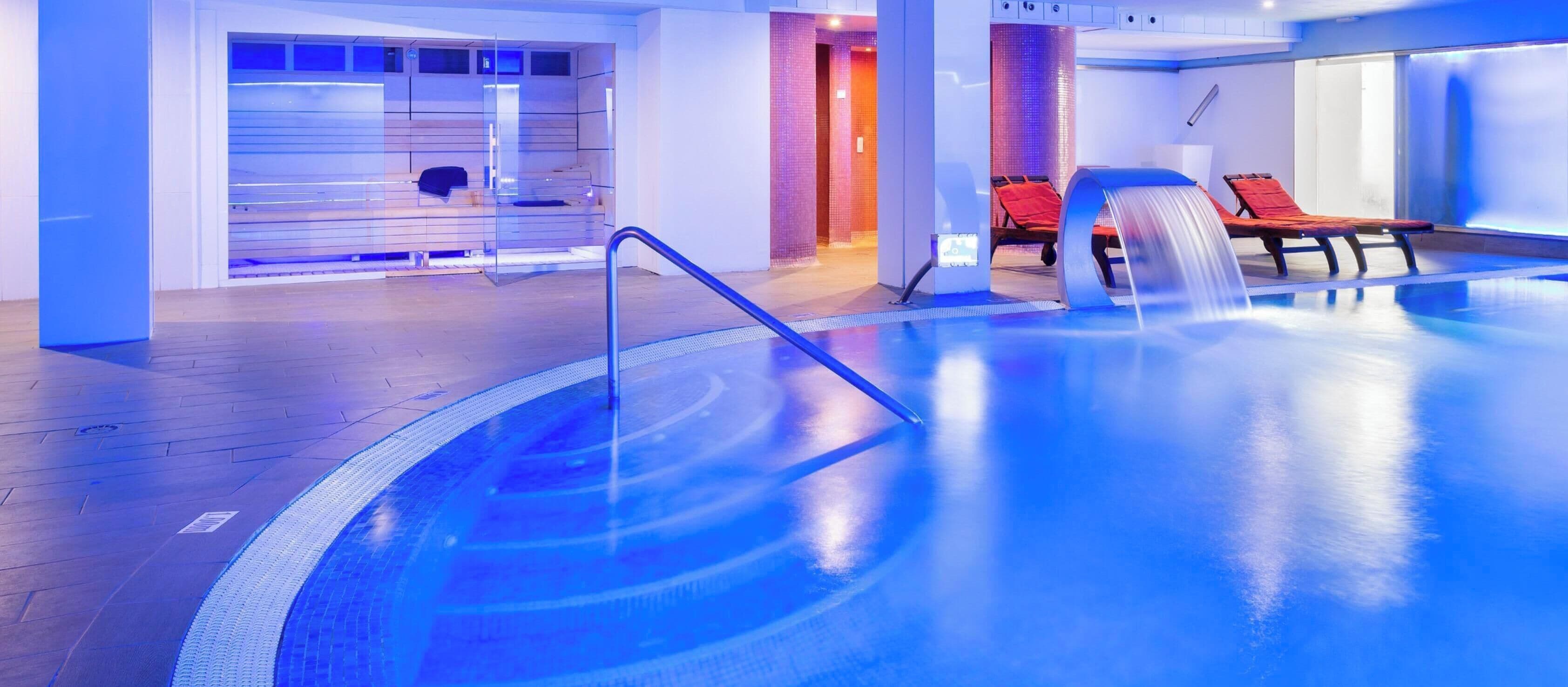 Une piscine intérieure moderne dotée d’un bassin à eau cristalline, de chaises longues et d’un espace spa clos par des parois de verre.