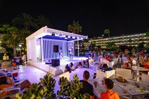 Evento al aire libre con escenario y público sentado en mesas, rodeado de palmeras y un edificio al fondo.