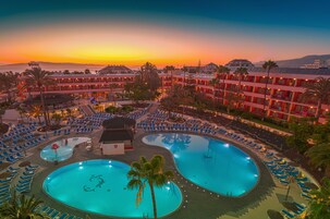 Um hotel com várias piscinas, um edifício grande com varandas e vista para o pôr do sol.