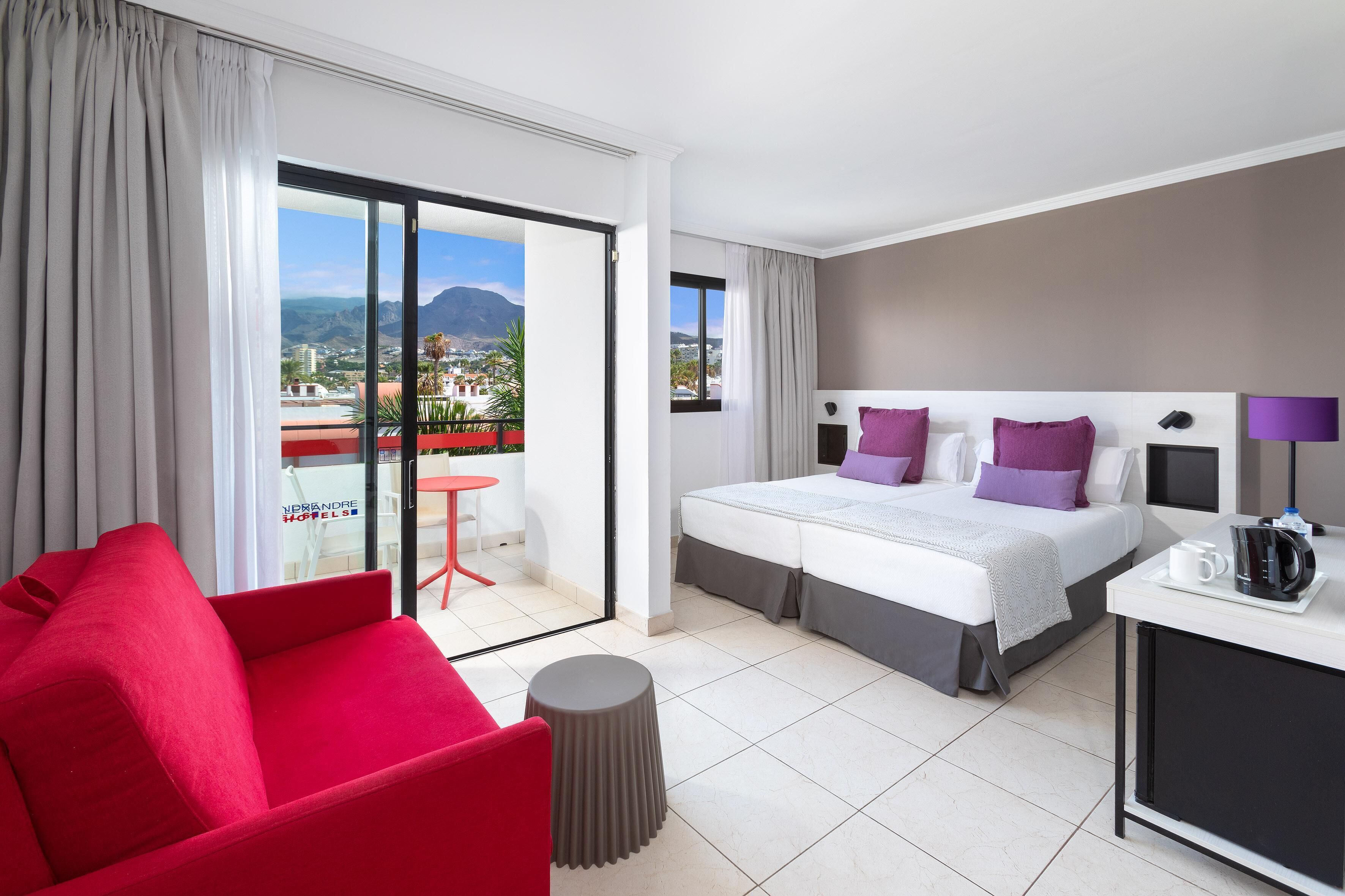 Habitación de hotel con cama, una silla roja, una mesita y vistas a la ciudad y las montañas.