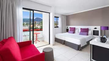 Habitación de hotel con cama, una silla roja, una mesita y vistas a la ciudad y las montañas.