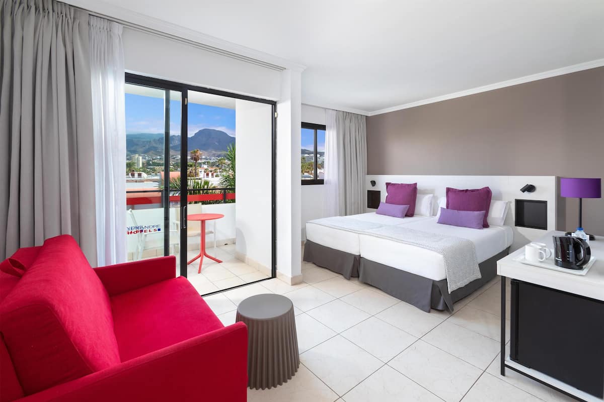 A hotel room with a bed, a red chair, a small table, and a view of the city and mountains through a sliding door.