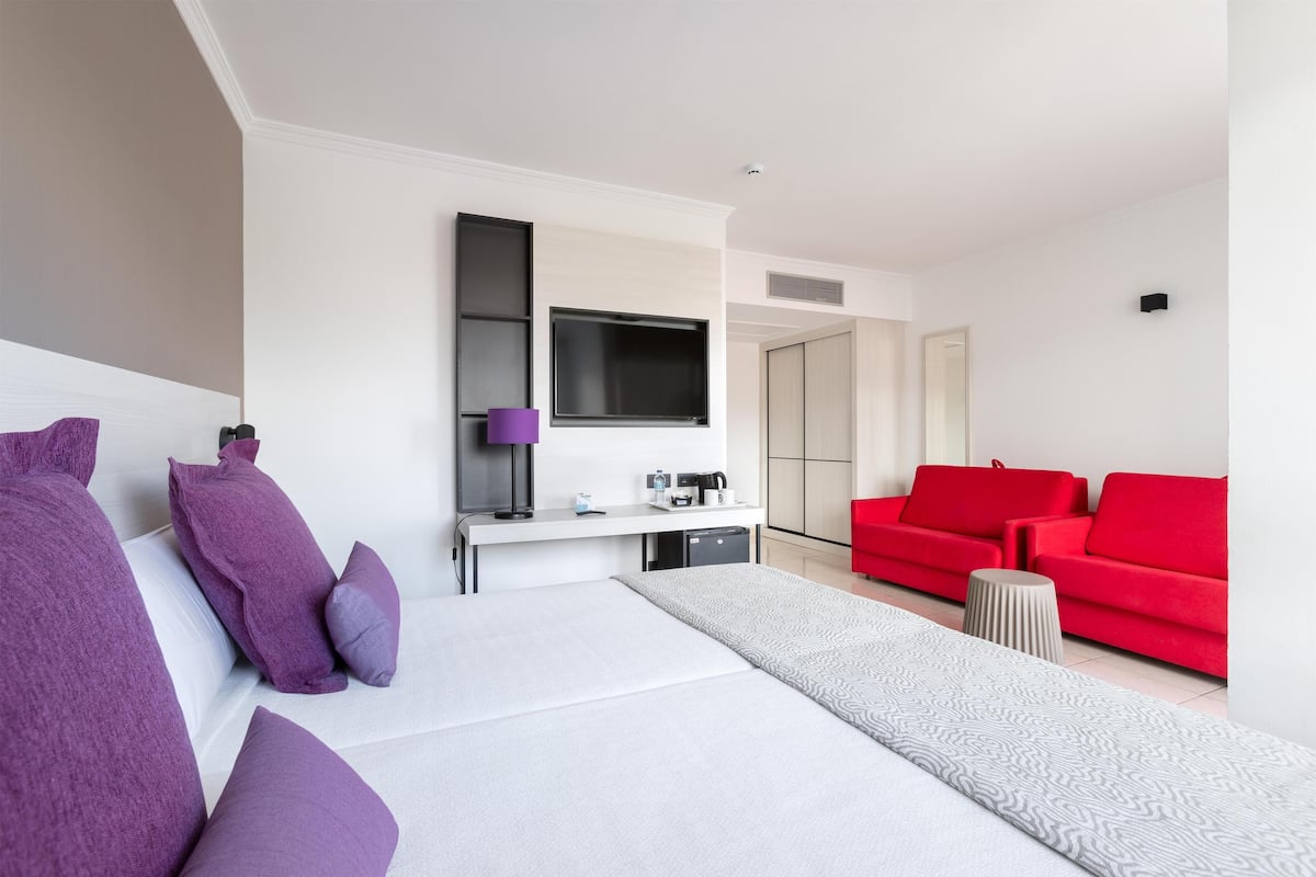 A modern hotel room with a large bed, a desk with a flat-screen TV, a red sofa, and a small side table.
