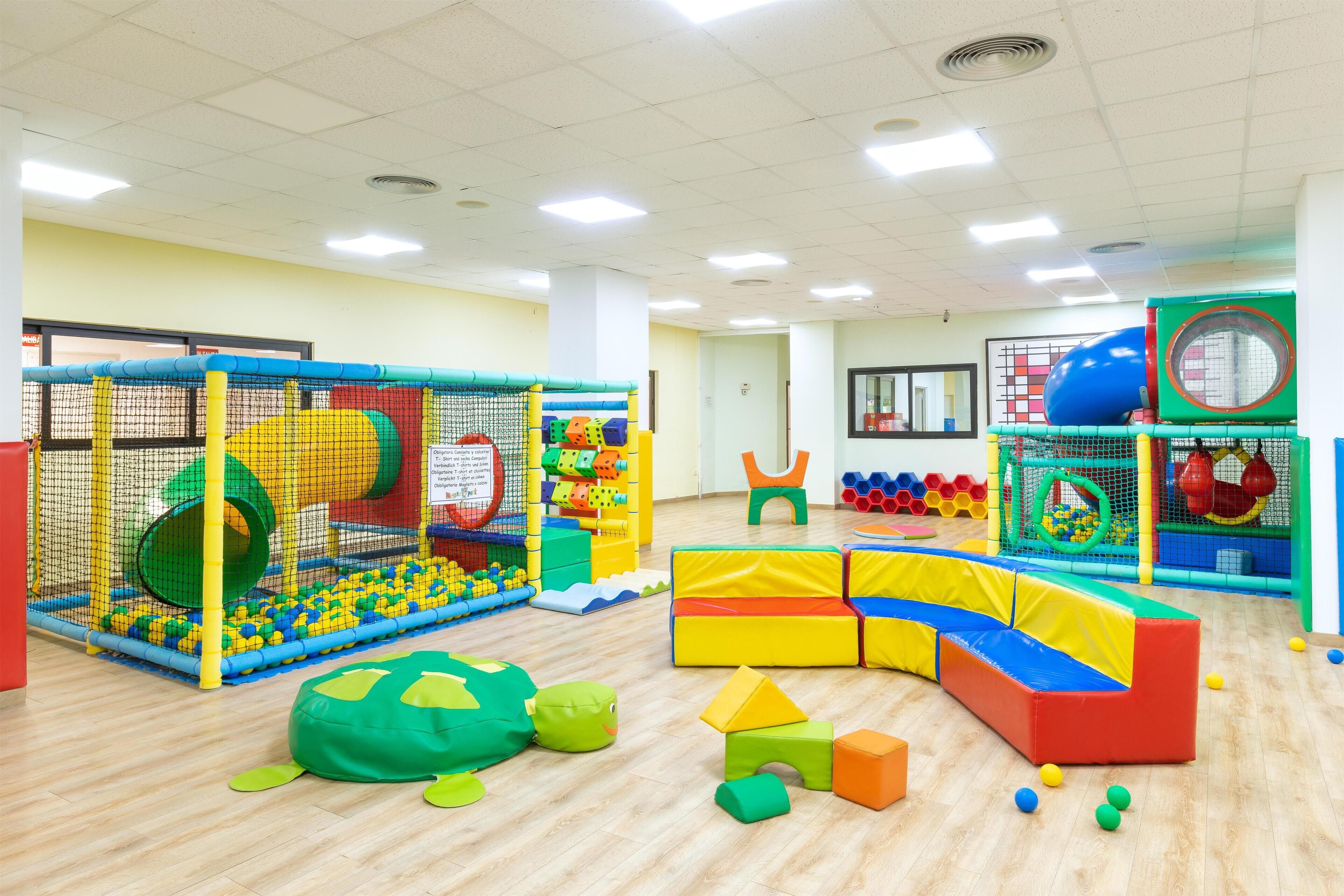 A brightly lit indoor play area with various play structures, including a ball pit, tunnels, and soft play equipment.