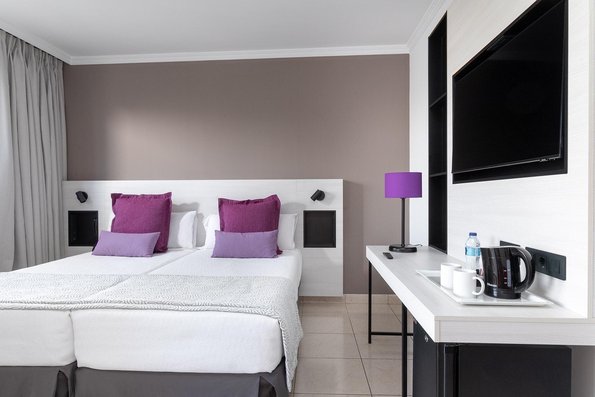 Quarto de hotel com cama de casal, escrivaninha com cafeteira e televisão.