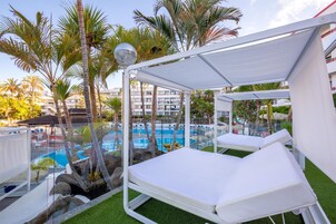Uma área à beira da piscina com espreguiçadeiras, um espaço coberto para sentar e vista para a piscina e palmeiras ao redor.