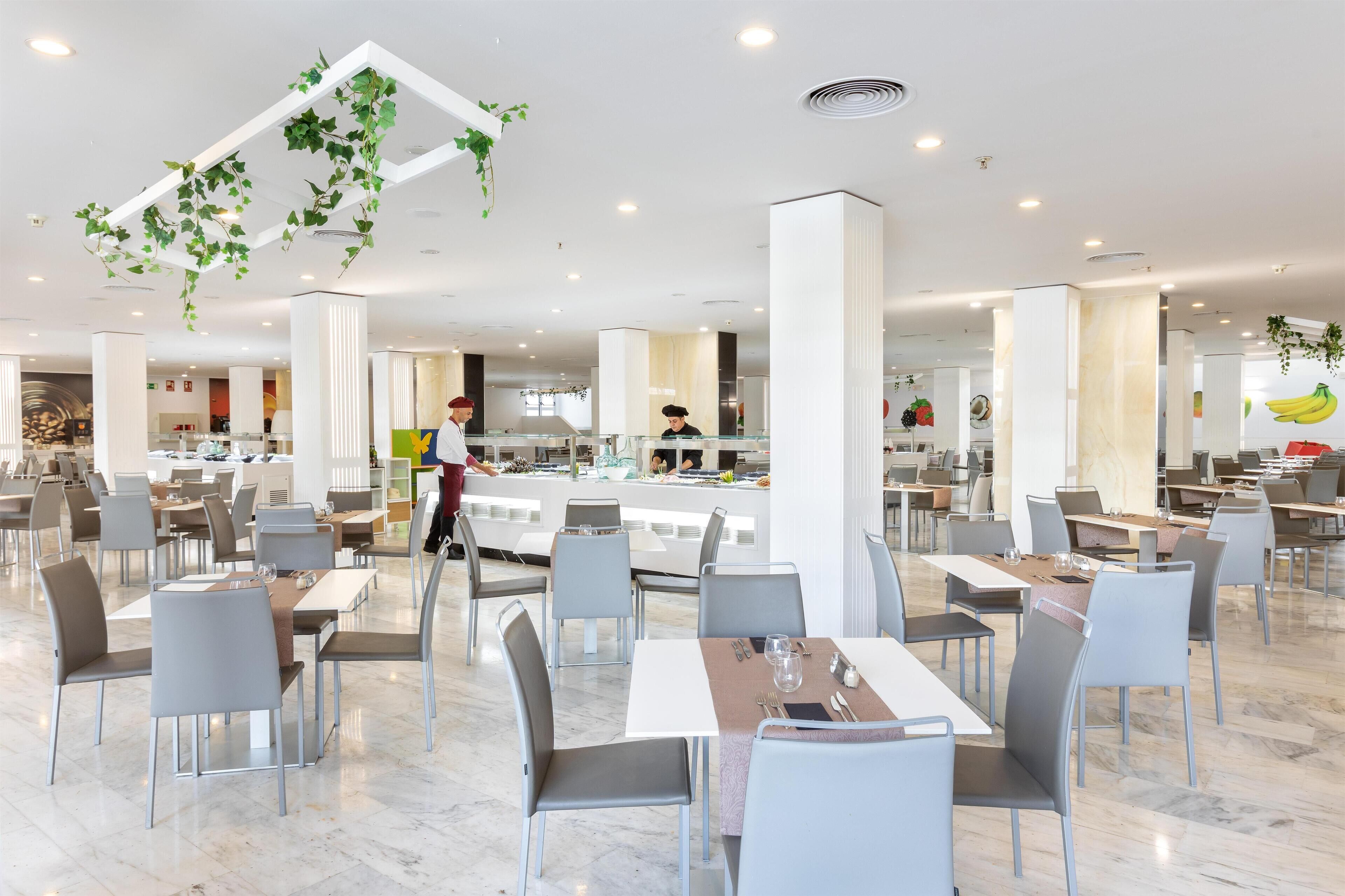 Un comedor amplio con varias mesas y sillas, una barra central y plantas colgantes.