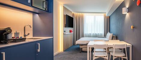 Una habitación de hotel compacta con kitchenette, una cama, zona de comedor y televisor montado en la pared.