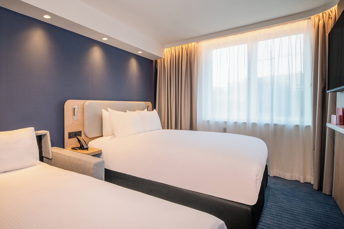 A hotel room with two beds, a nightstand, a desk, and a large window with curtains.