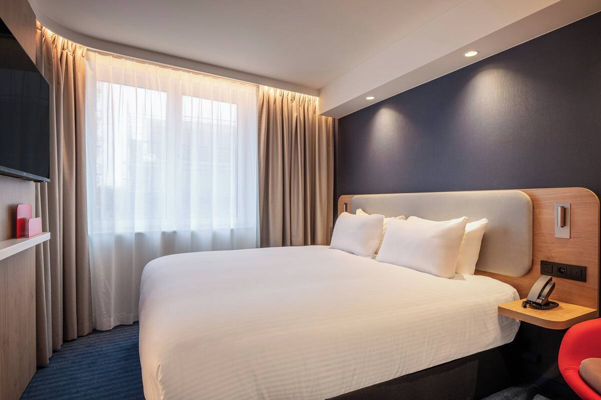 A hotel room with a large bed, a bedside table with a phone, a red chair, and a TV on a shelf.