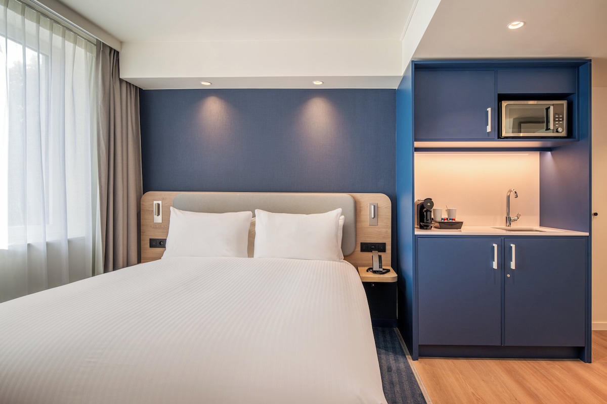 A hotel room with a large bed, a small kitchenette with a microwave, and a nightstand with a phone and a cup.