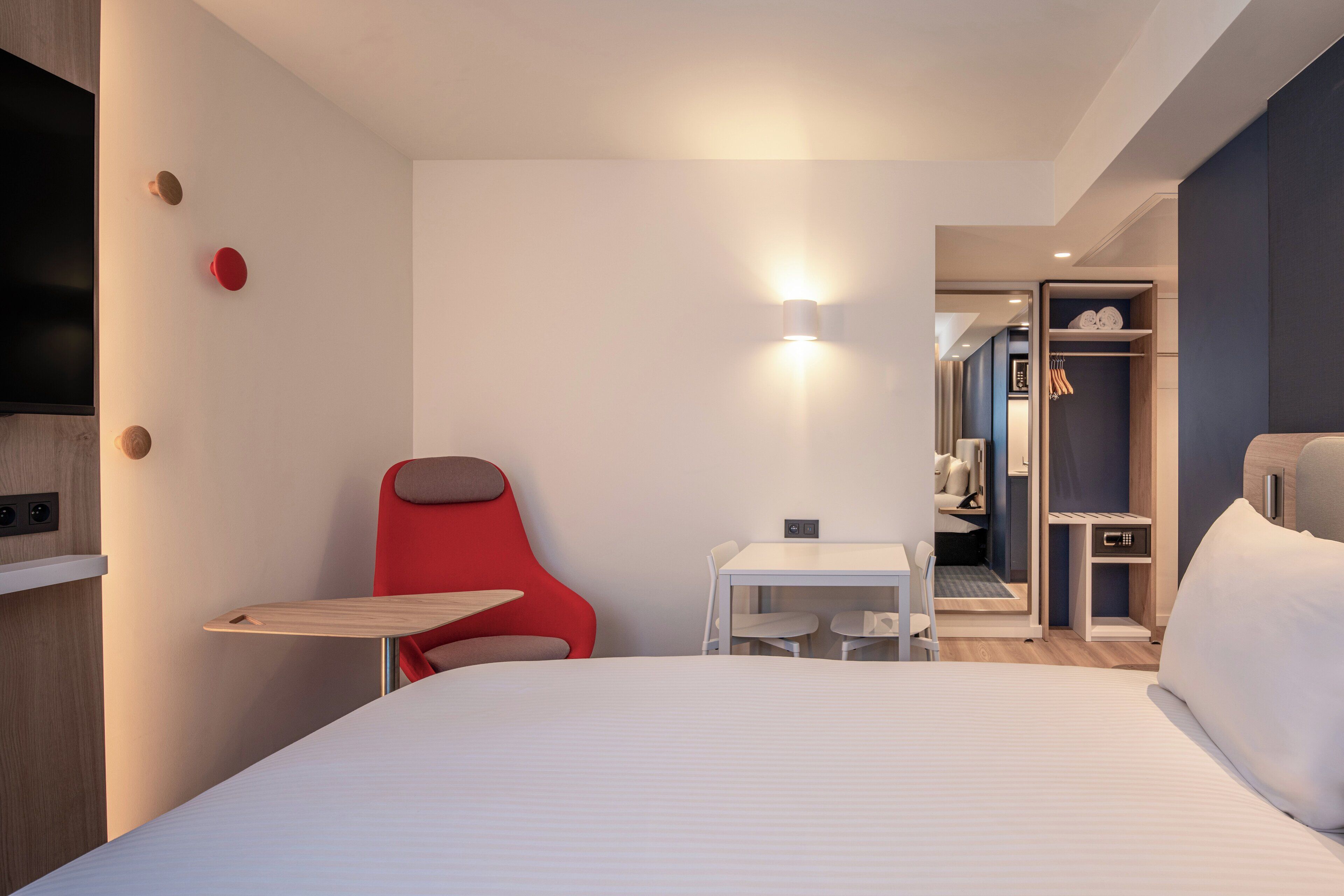 A modern hotel room with a bed, a small table, a red chair, a TV, and a wardrobe.
