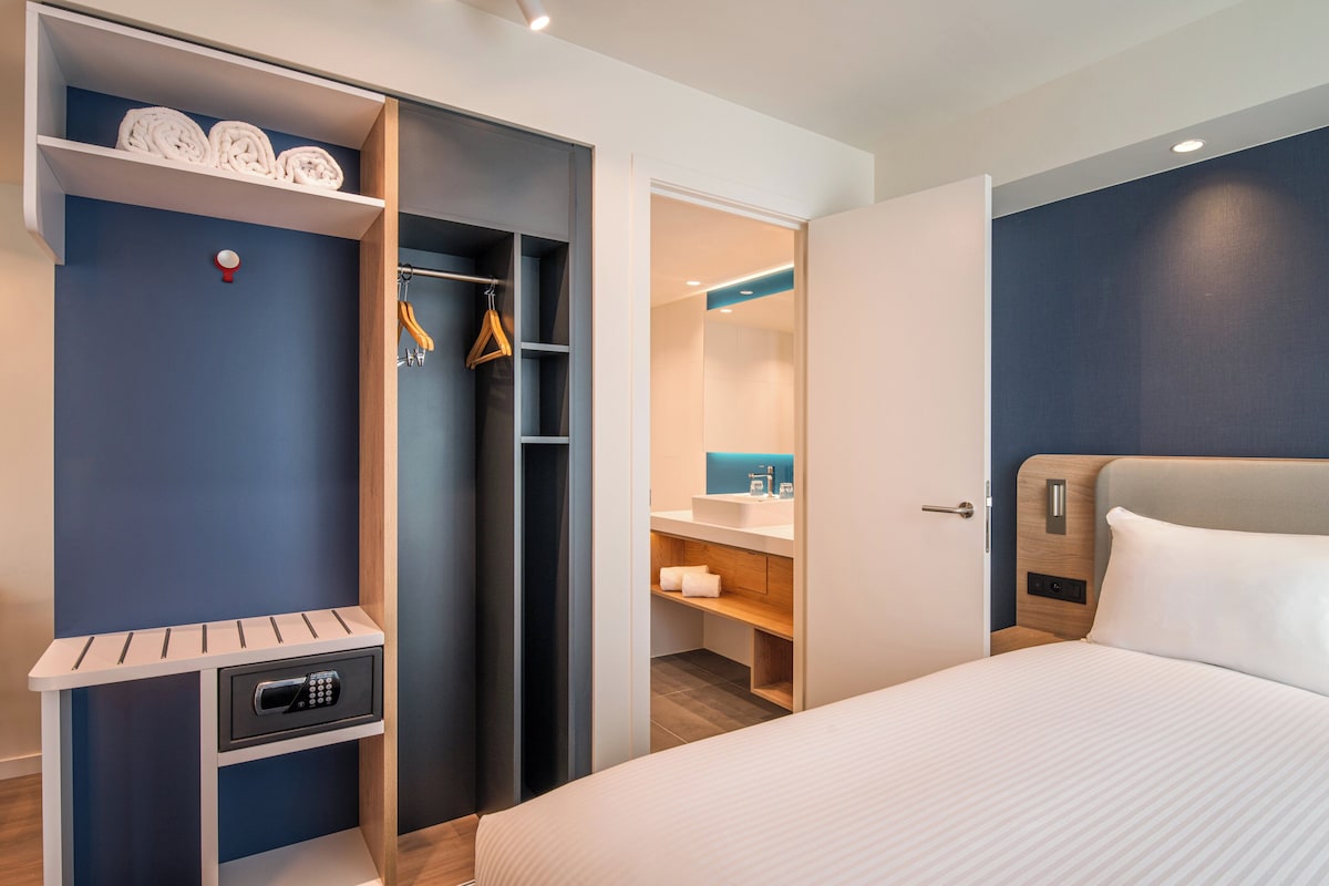 A hotel room with a bed, a walk-in closet with shelves, a small bathroom, and a bedside table with a lamp.
