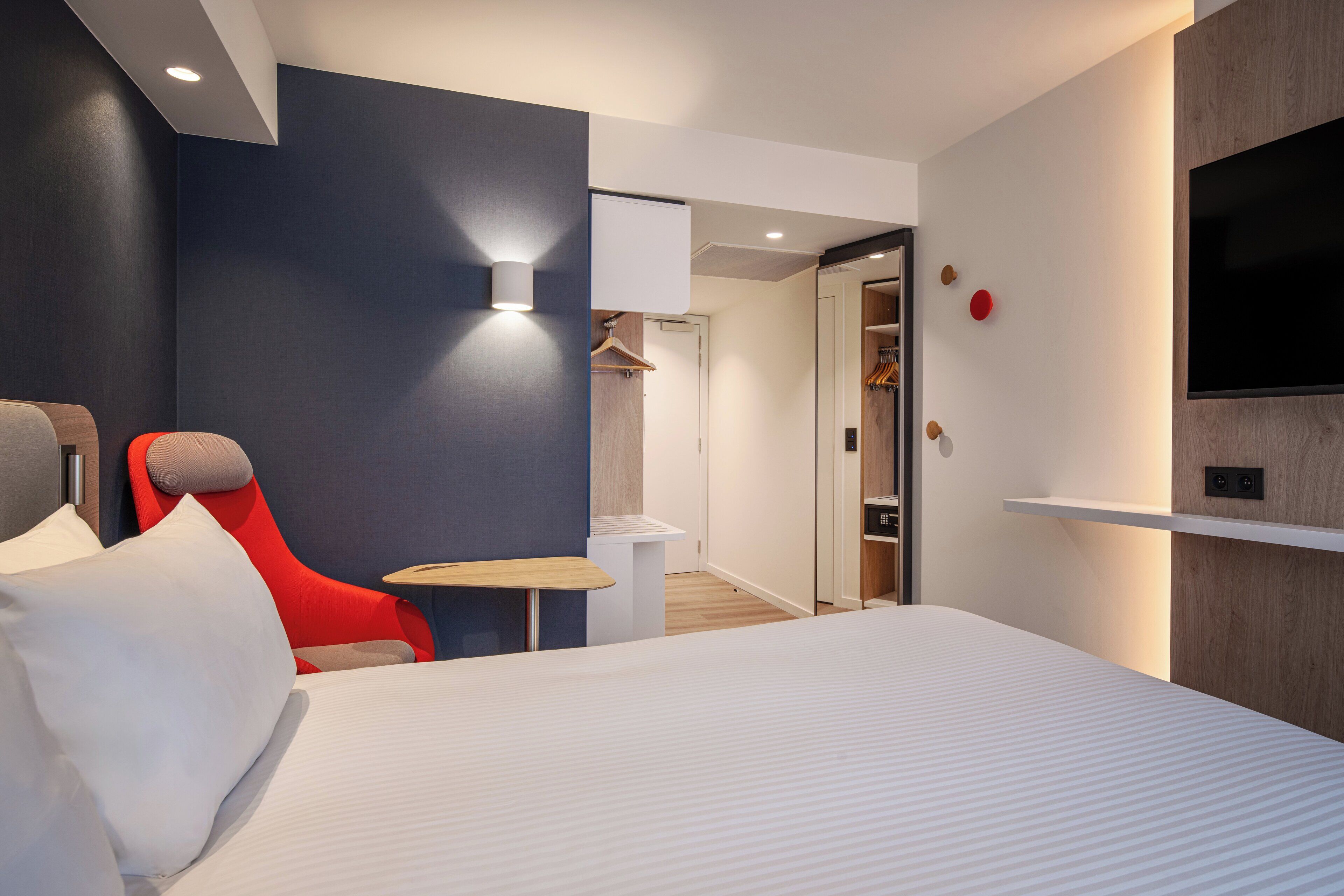 Et moderne hotelværelse med en stor seng, en rød lænestol, et lille bord og et fjernsyn ophængt på væggen.