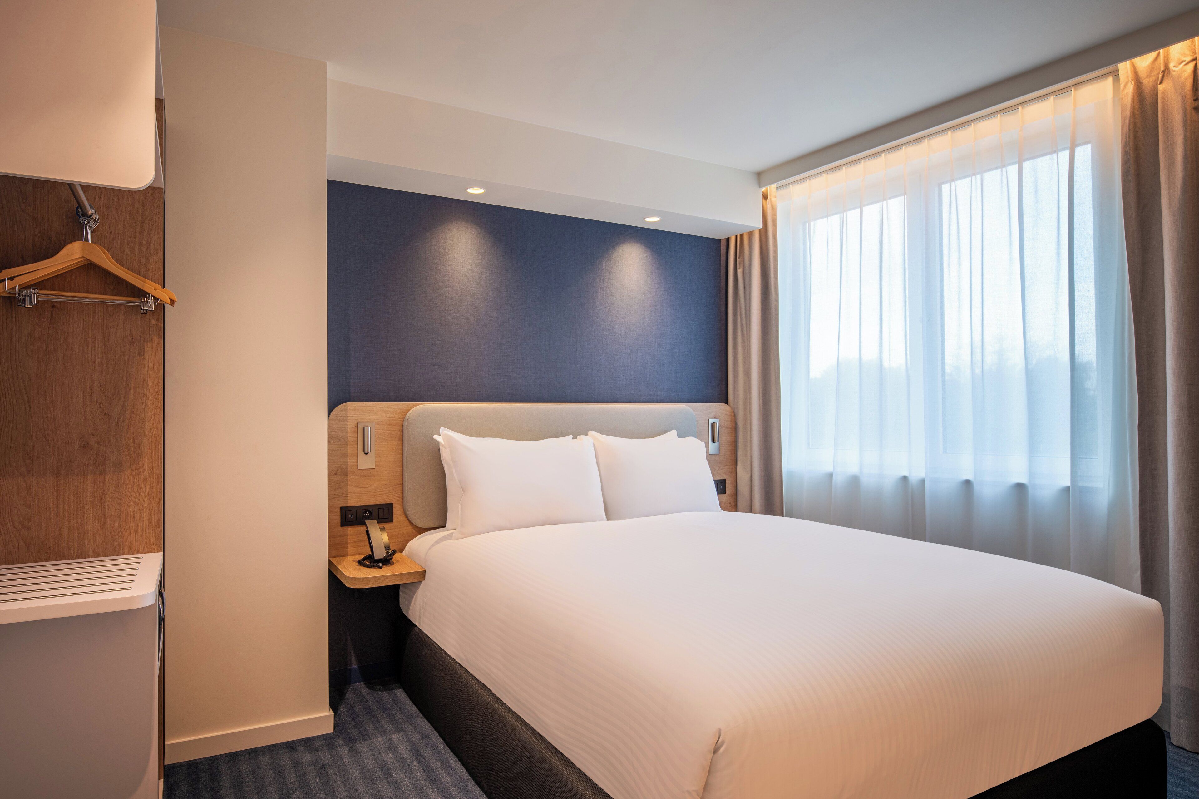 A hotel room with a large bed, a wooden wardrobe, a nightstand, and a window with curtains.