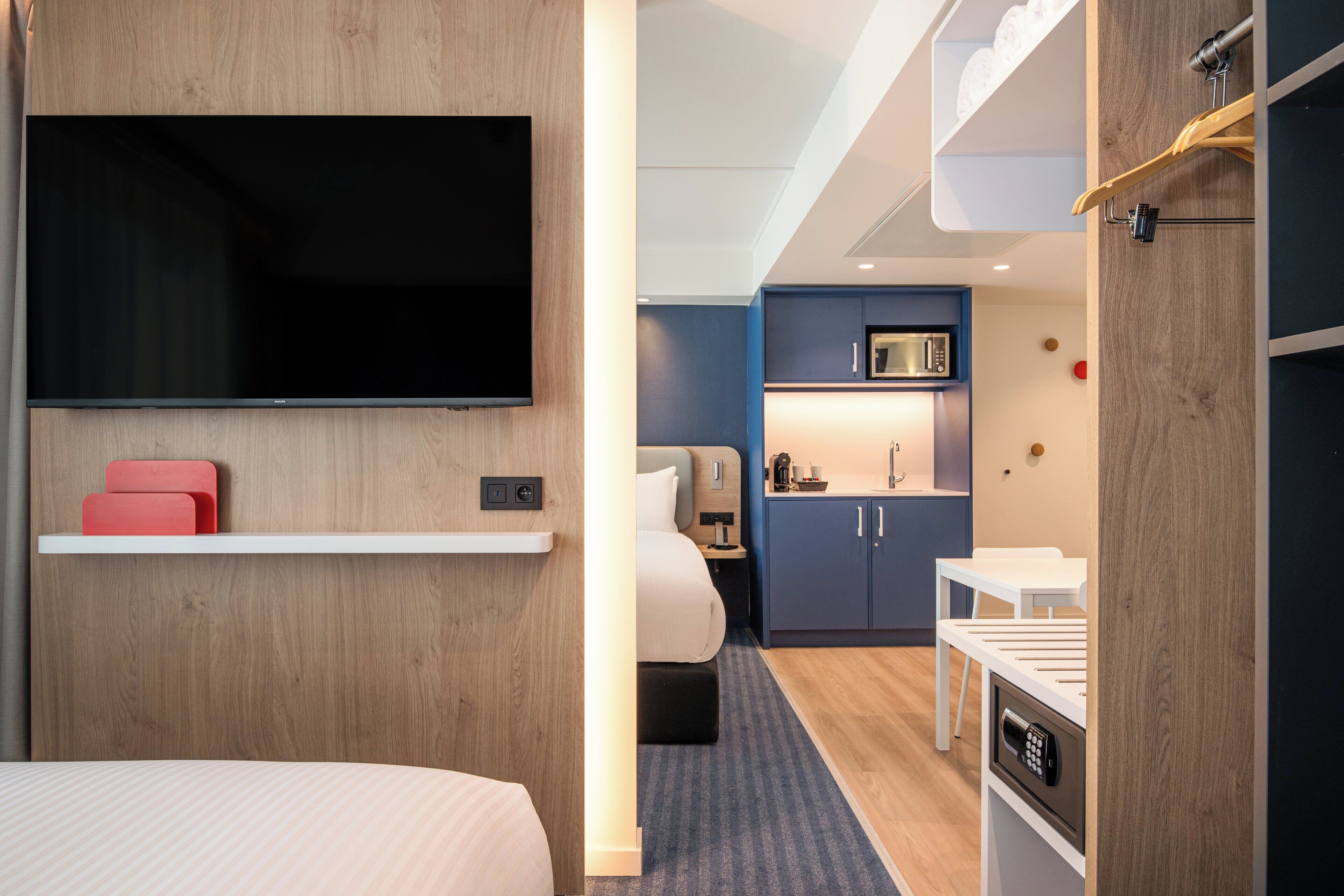 A hotel room with a bed, a TV mounted on the wall, a small kitchenette, and a desk with a chair.