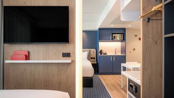 A hotel room with a bed, a TV mounted on the wall, a small kitchenette, and a desk with a chair.