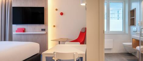 Una habitación de hotel moderna con una cama, un televisor montado en la pared, una mesita con una silla y un baño con inodoro y lavabo.