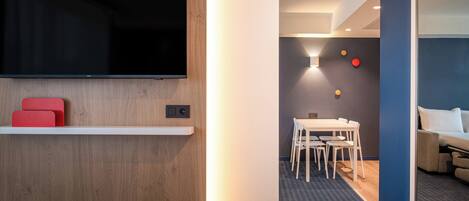 A hotel room with a flat-screen TV mounted on the wall, a small shelf with red cushions, a white bed, a dining area with a table and chairs, and a blue accent wall.