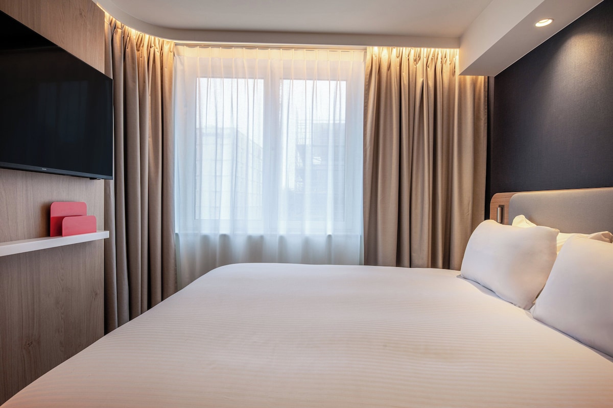 A modern hotel room with a large bed, a flat-screen TV mounted on the wall, a bedside table with a red object, and a window with sheer curtains.