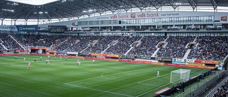 Un estadio con múltiples niveles de asientos, un amplio campo de juego y publicidad para VDK Bank y Play Sports.