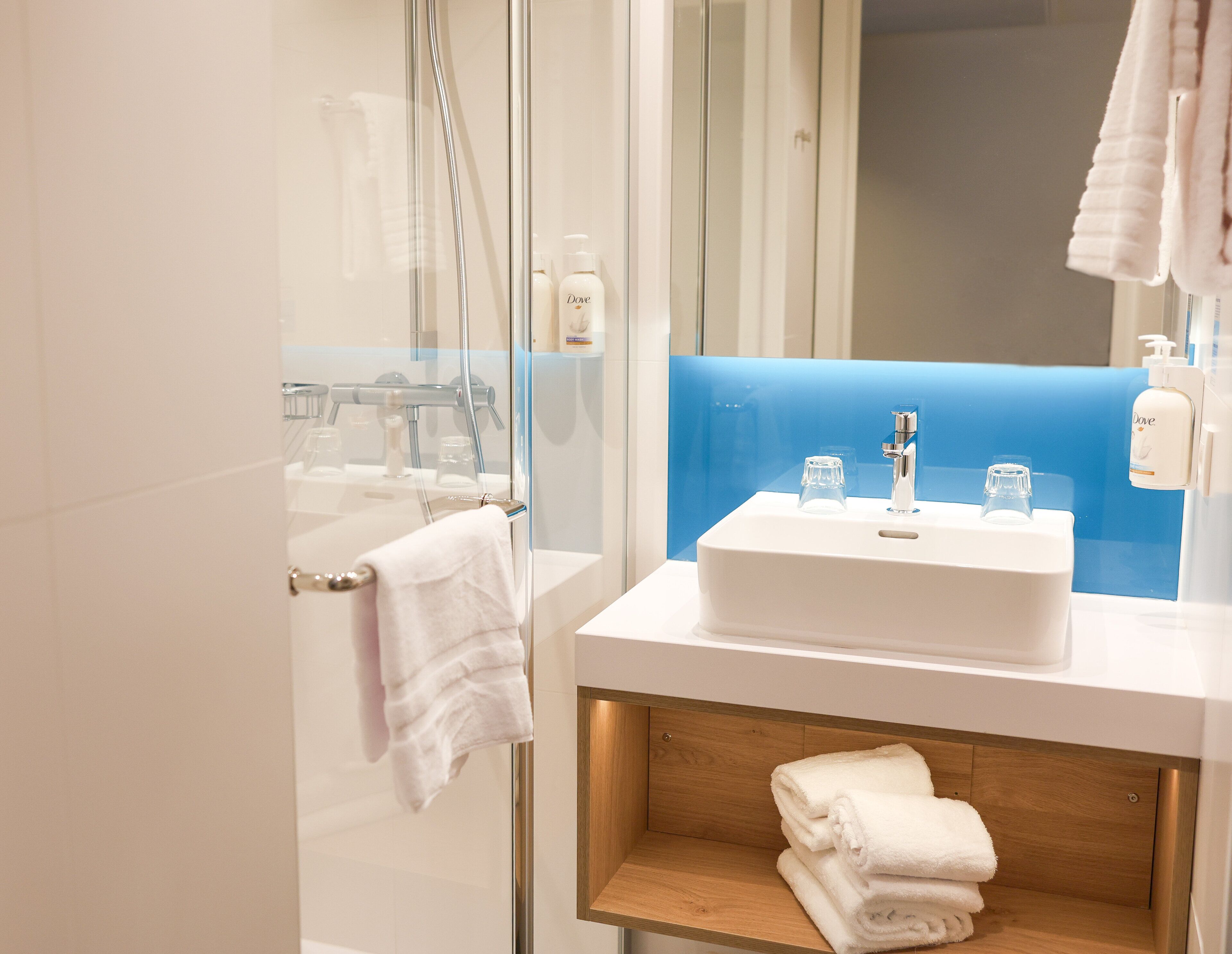 Un baño moderno con cabina de ducha de vidrio, un lavabo blanco con estante de madera y una pared de acento azul.