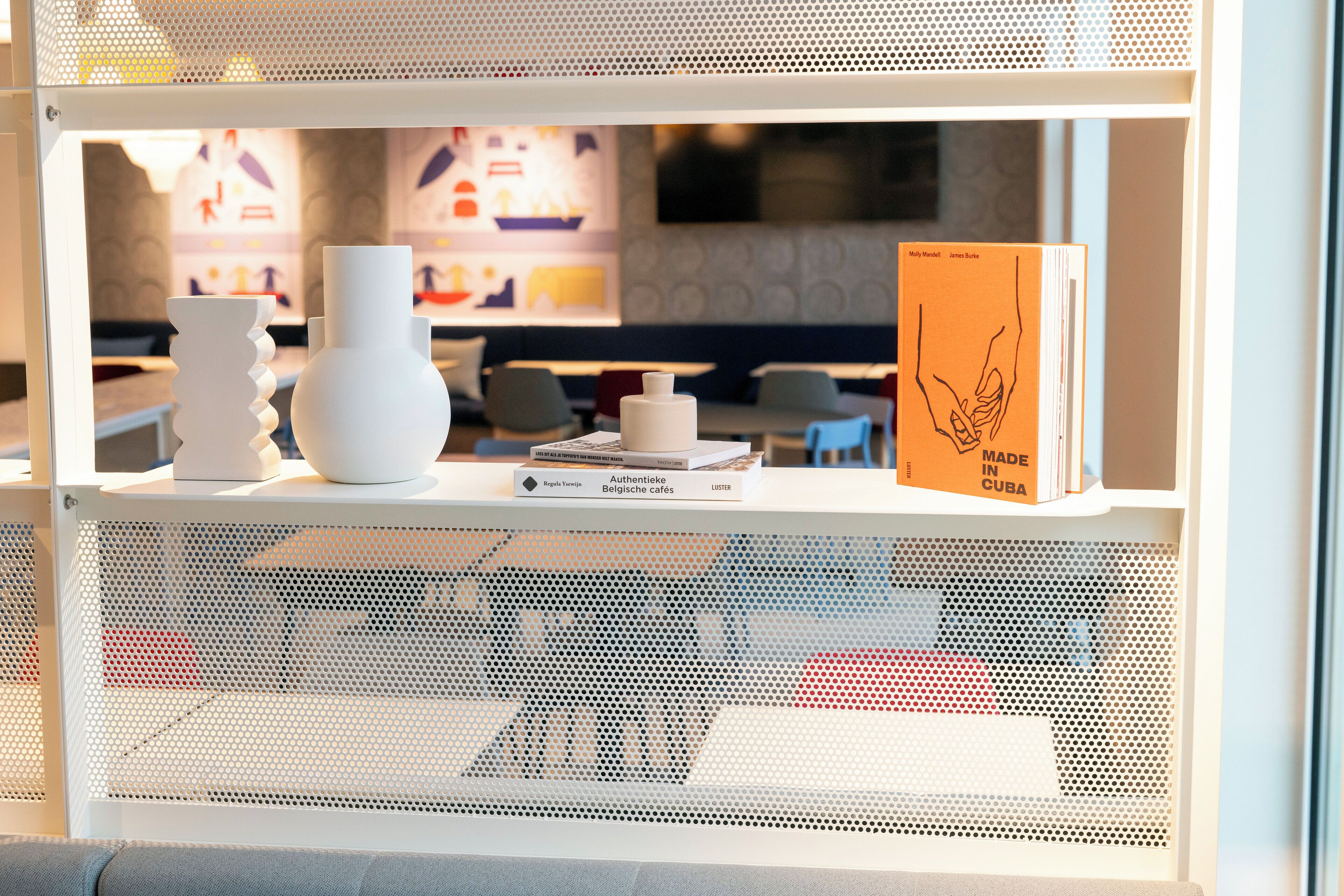 Estante de exhibición con jarrones decorativos y un libro titulado 'Authentieke Belgische cafes' en un ambiente de cafetería.