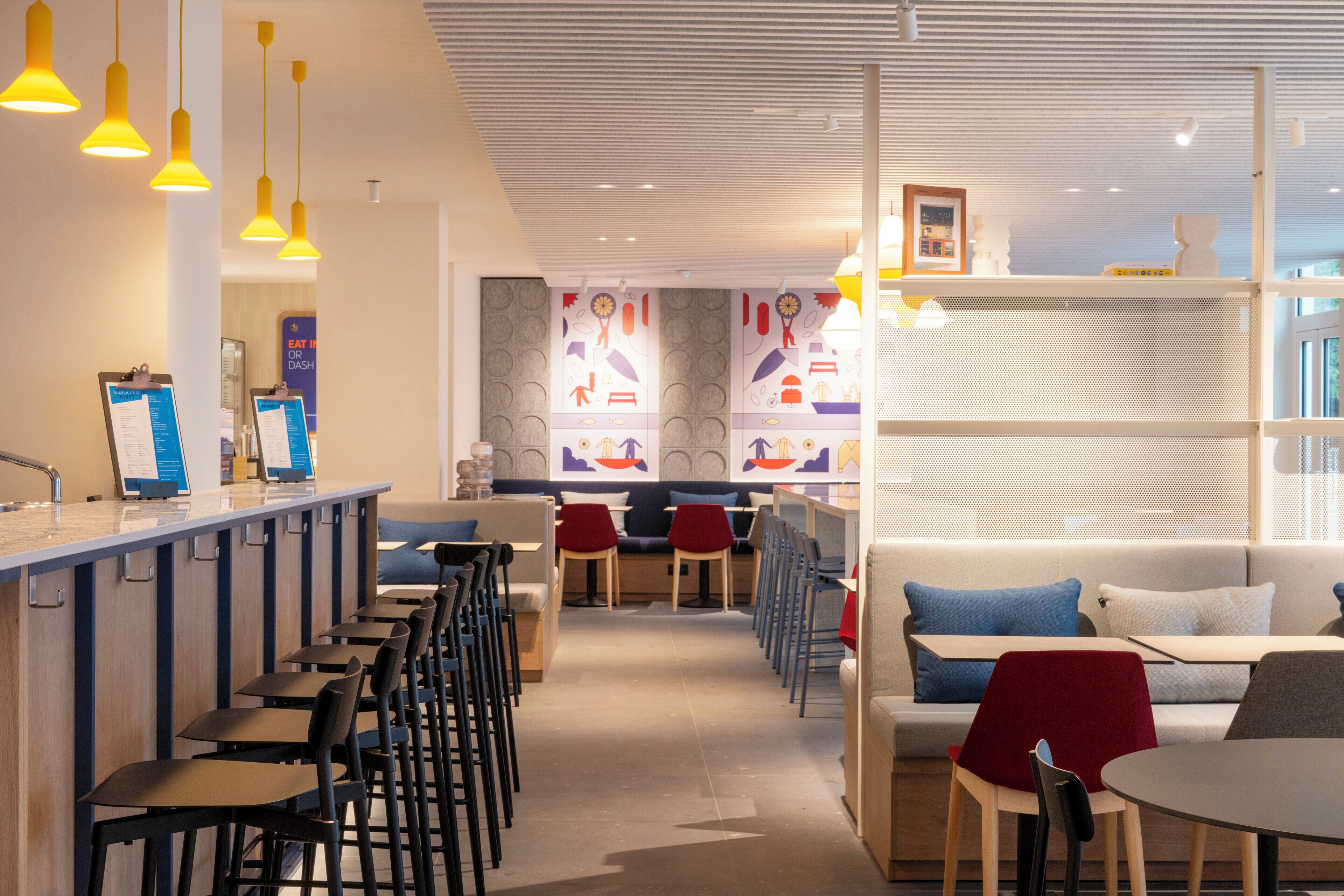 Una cafetería moderna con una barra larga, varias zonas de estar y una pared decorada al fondo.