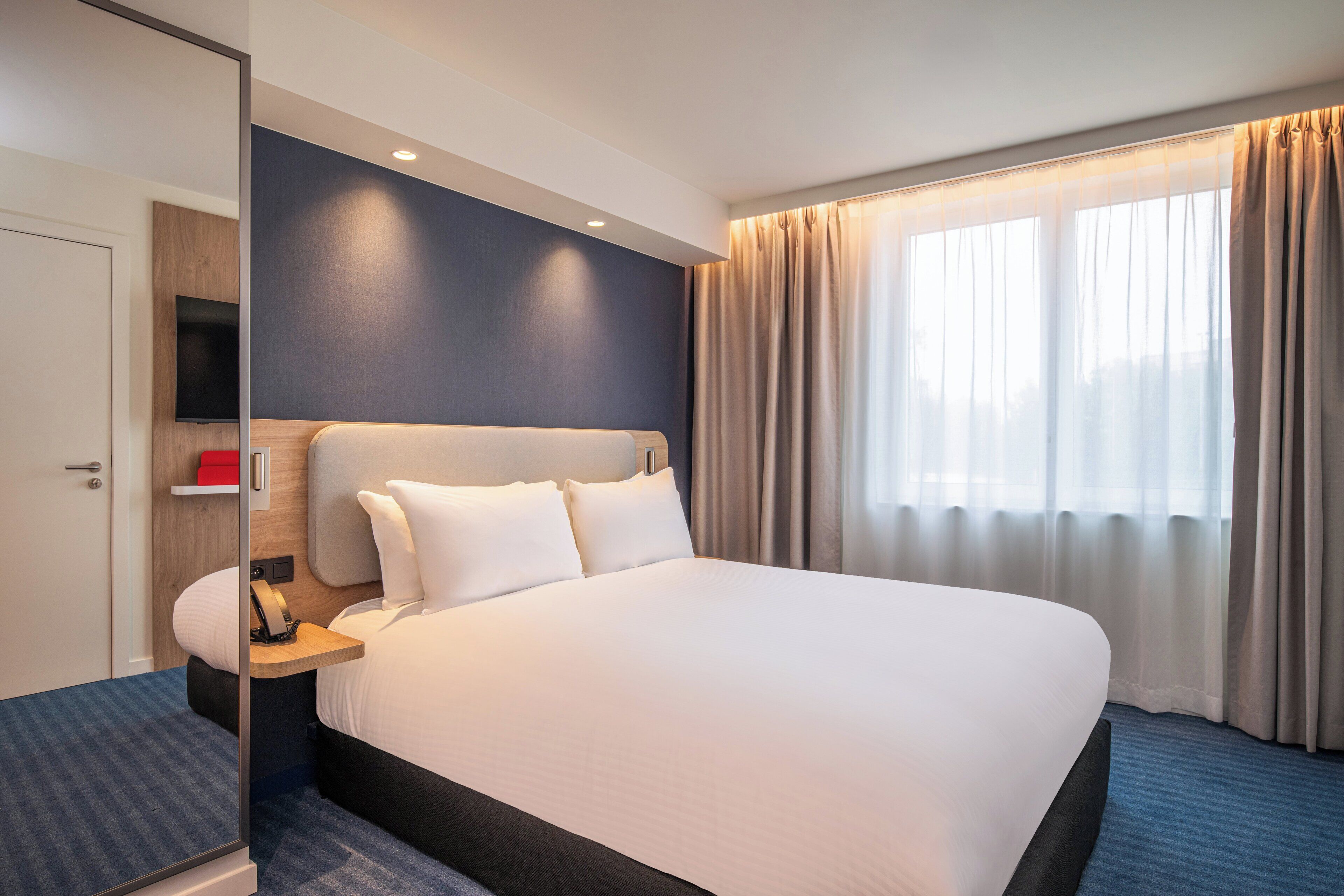 Una habitación de hotel moderna con una cama grande, una mesita de noche, un televisor de pantalla plana y una ventana con cortinas.