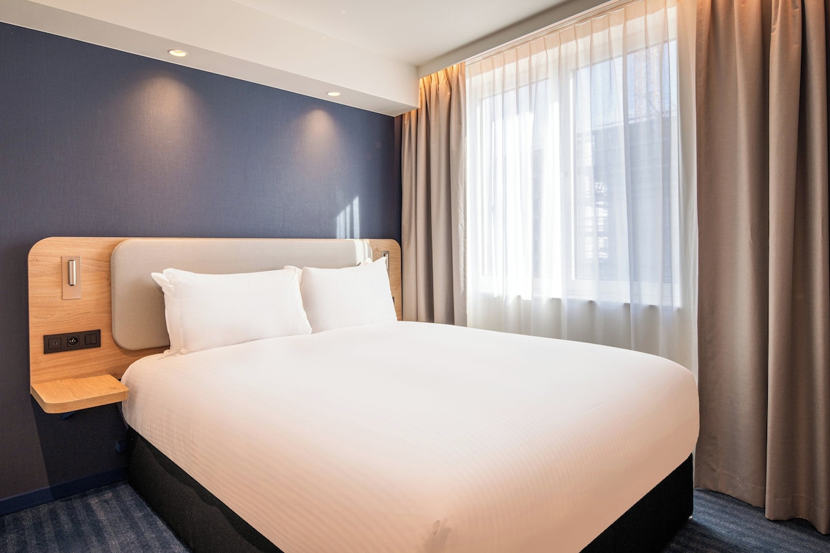 A modern hotel room with a large bed, a wooden headboard, a bedside table, and a window with curtains.