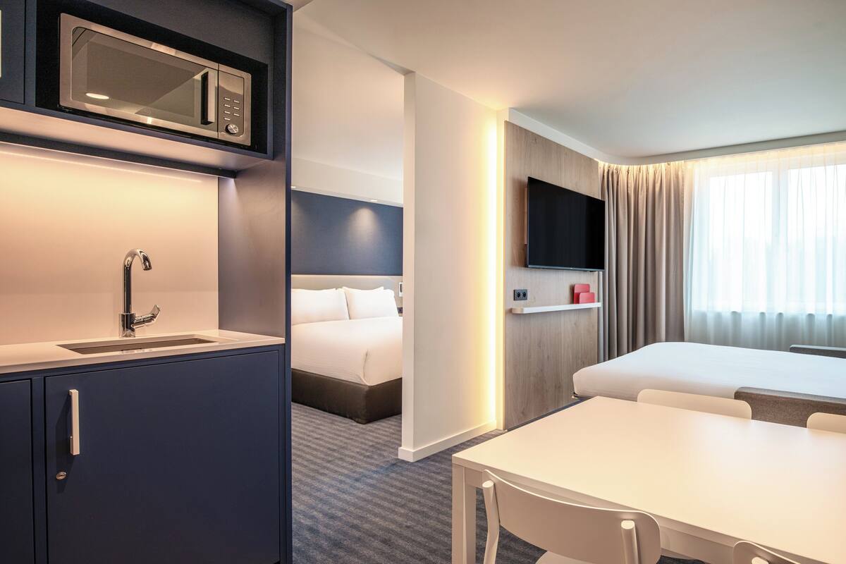 A hotel room with a kitchenette, a bed, a TV, and a window with curtains.