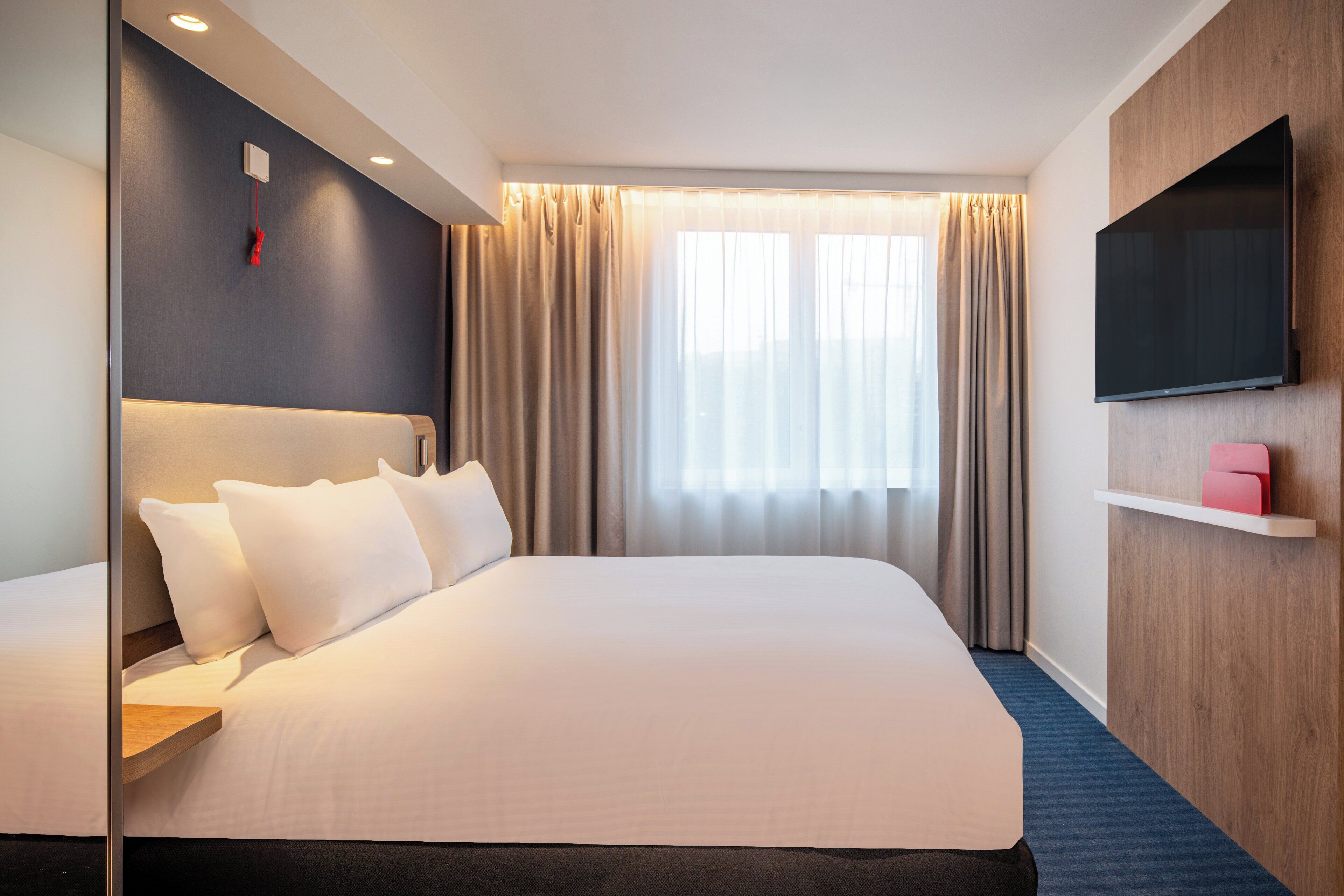 A hotel room with a large bed, a flat-screen TV mounted on the wall, and a window with curtains.