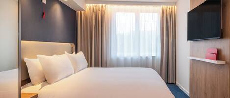 A hotel room with a large bed, a flat-screen TV mounted on the wall, and a window with curtains.