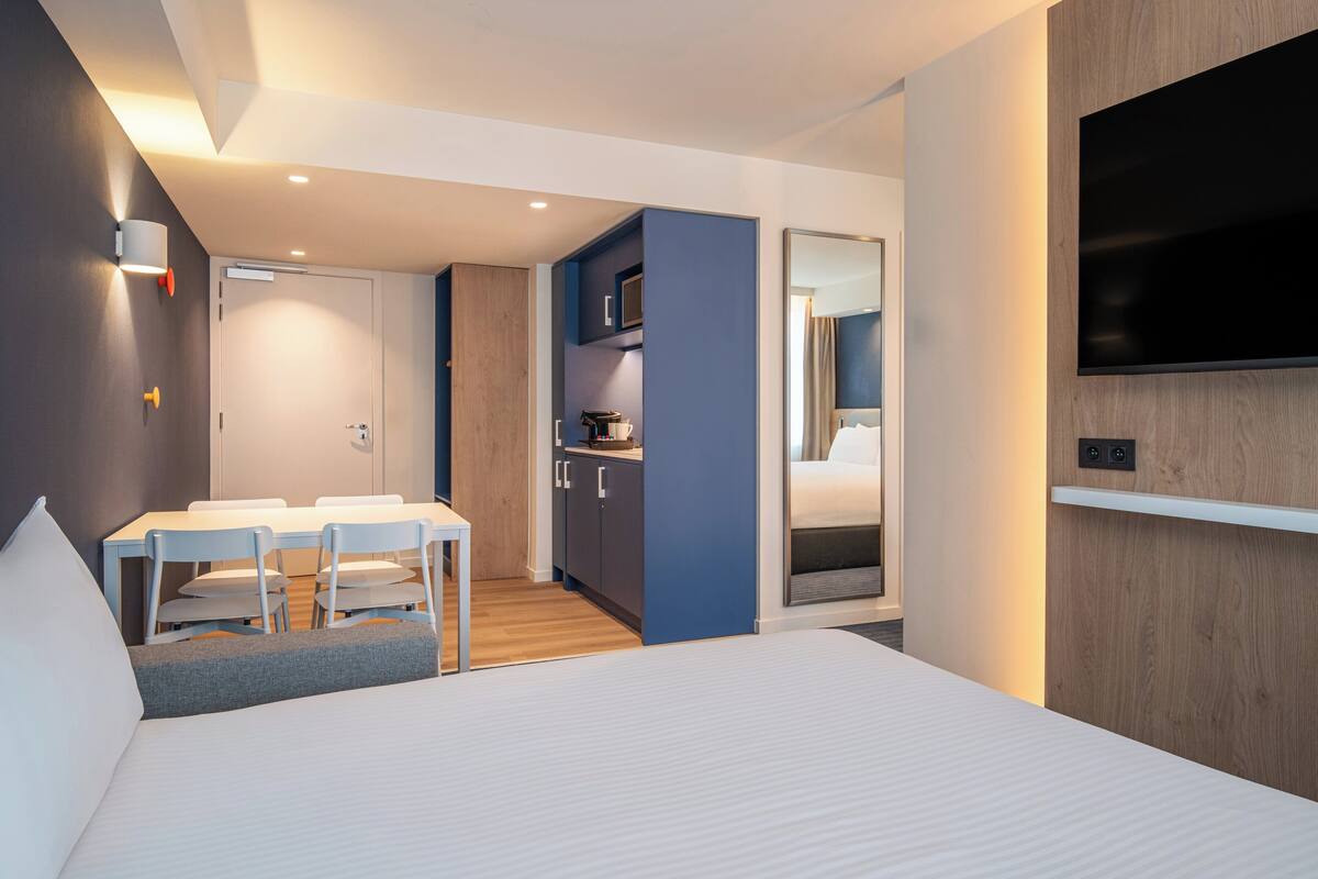 A modern hotel room with a bed, a dining area with chairs, a kitchenette, and a TV mounted on the wall.