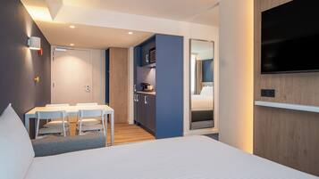 Una habitación de hotel moderna con una cama, un rincón de comedor con sillas, una kitchenette y un televisor montado en la pared.