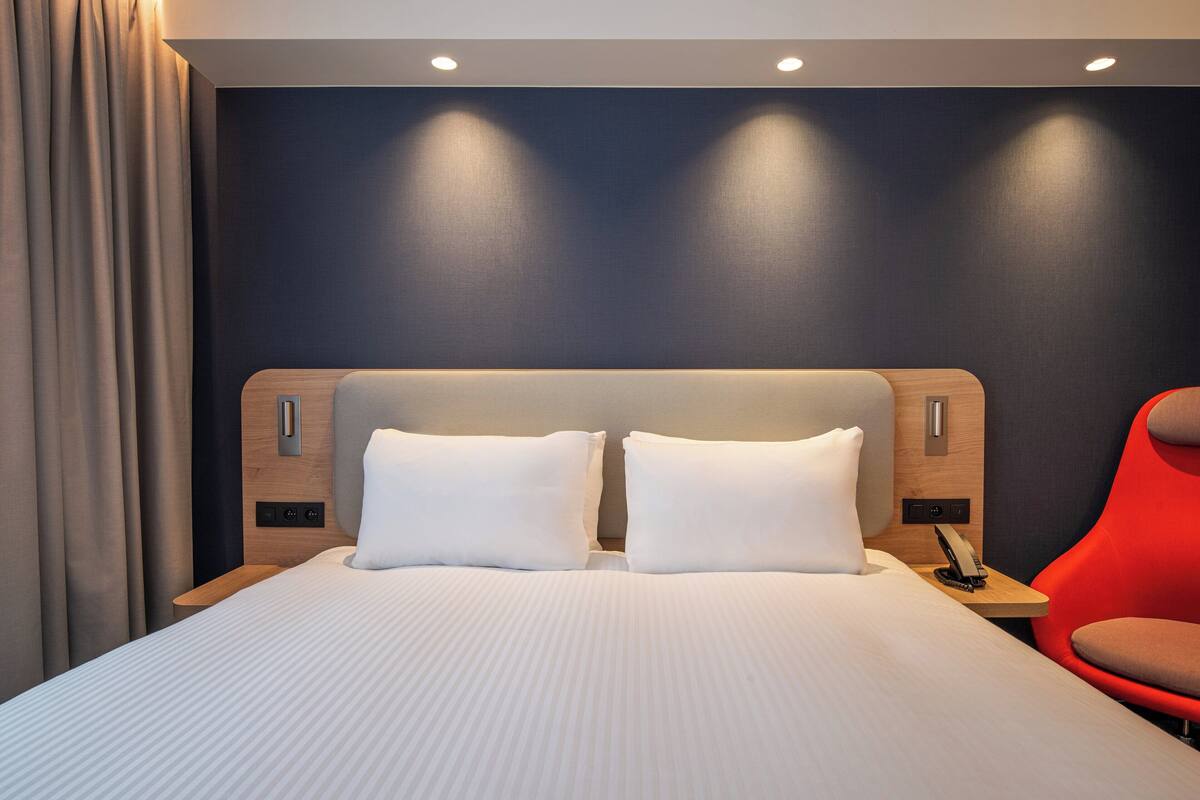 A modern hotel room with a large bed, a wooden headboard, a red chair, and a dark wall.