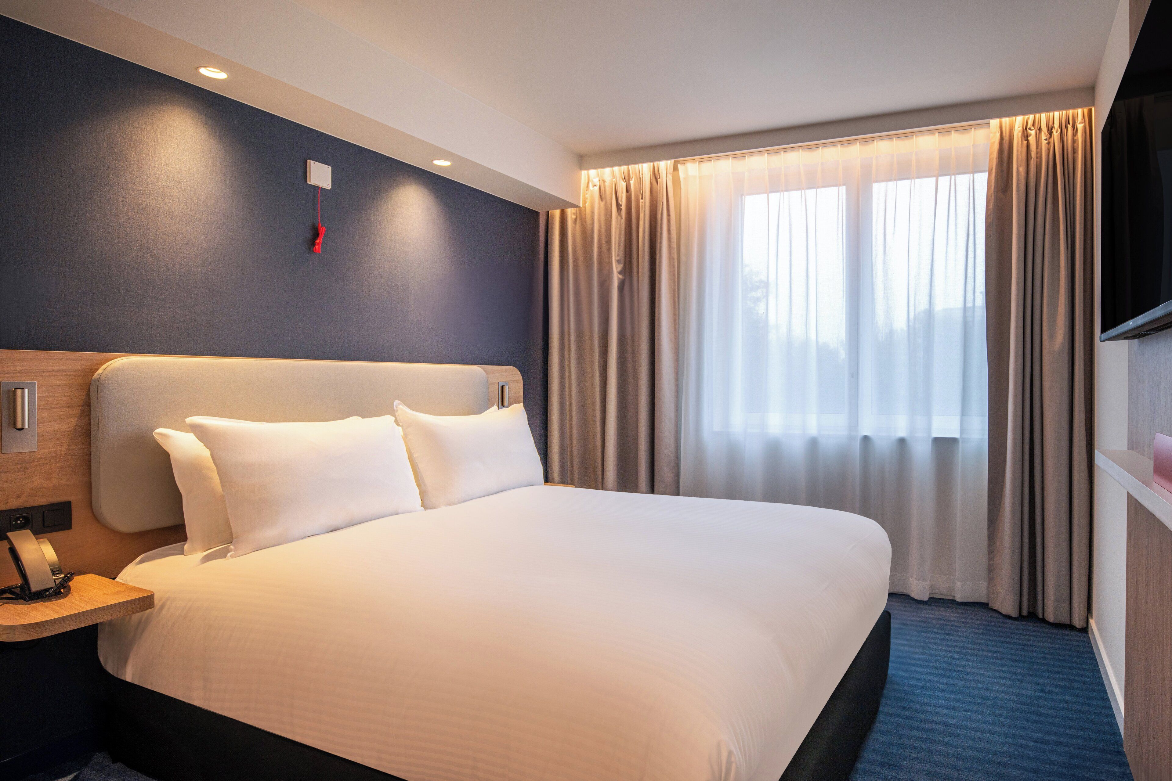 A hotel room with a large bed, a bedside table with a lamp, a flat-screen TV mounted on the wall, and a window with curtains.
