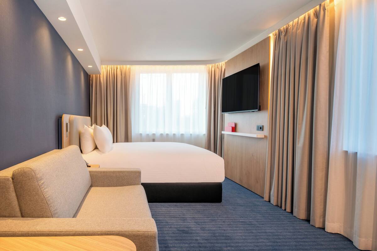 A modern hotel room with a large bed, a sofa, a flat-screen TV mounted on the wall, and a window with curtains.
