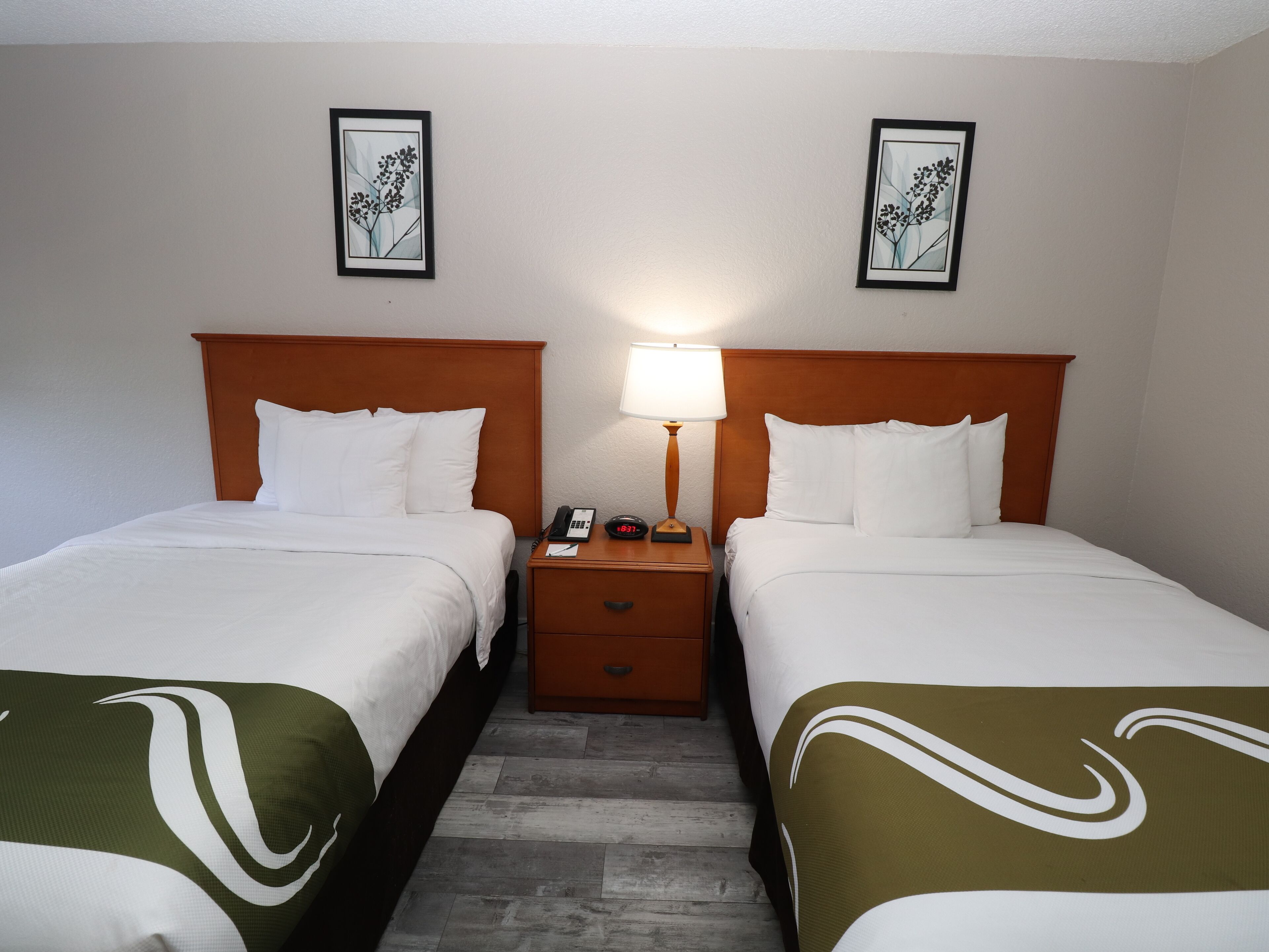 A hotel room with two beds, a nightstand with a lamp, and framed artwork on the wall.