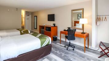 Habitación de hotel con cama, escritorio, silla, televisión, espejo y perchero con toallas.