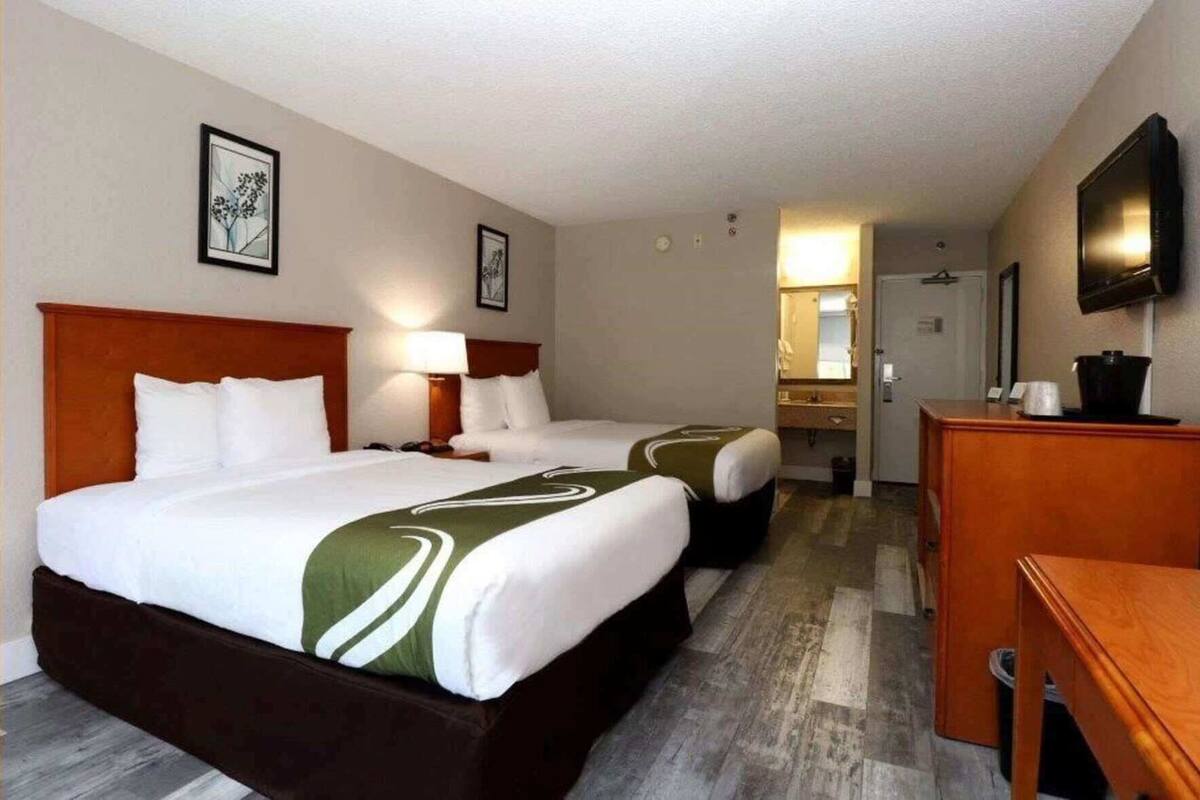 Hotellihuone, jossa on kaksi sänkyä, työpöytä, televisio ja kylpyhuone, joka näkyy avoimesta ovesta.