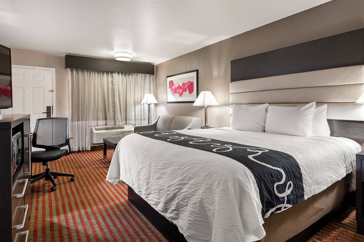 A hotel room with a large bed, two bedside lamps, a desk with a chair, a flat-screen TV, and a window with curtains.