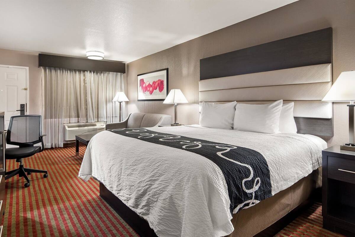 A hotel room with a bed, desk, chair, lamps, and a window with curtains.